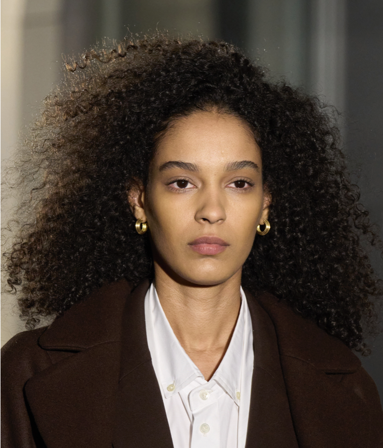 A person with curly hair wearing a brown jacket and gold earringsDescription automatically generated