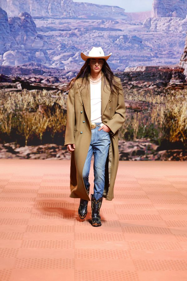 A person wearing a cowboy hat and long coat walking on a runway Description automatically generated